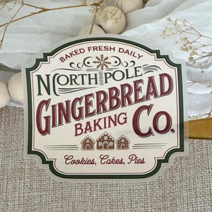 May include: A white and green sign with the text "Baked Fresh Daily" and "North Pole Gingerbread Baking Co." in red and green lettering. The sign also features a snowflake and three gingerbread houses. The text "Cookies, Cakes, Pies" is at the bottom of the sign.