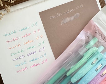 May include: A collection of colorful gel pens, labeled "SARASA CLIP" and "milk color 0.5", are displayed alongside handwritten color tests on paper. The pens are in shades of green, blue, purple, and pink. The text "WALDEN WOODWORKERS ISTANBUL" is visible.