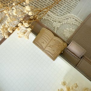 Puede incluir: Un diario de cuero beige con un cierre dorado en forma de libro con un diseño de pluma y estrellas. El diario está abierto en una página en blanco con líneas de cuadrícula, con flores secas y detalles de encaje.