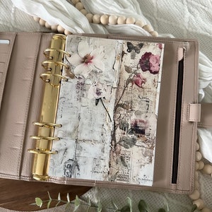 May include: A blush pink leather planner with gold rings and a floral patterned insert. The insert features a white flower, a butterfly, and a pink flower with green leaves.