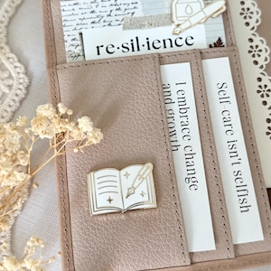 Op de afbeelding: Een lichtbruine leren planner met een goudkleurige boek- en penpin. De planner bevat kaarten met de woorden "re-sil·ience", "I embrace change and grow" en "Self-care isn't selfish". Decoratieve elementen zijn een kanten rand en droogbloemen.