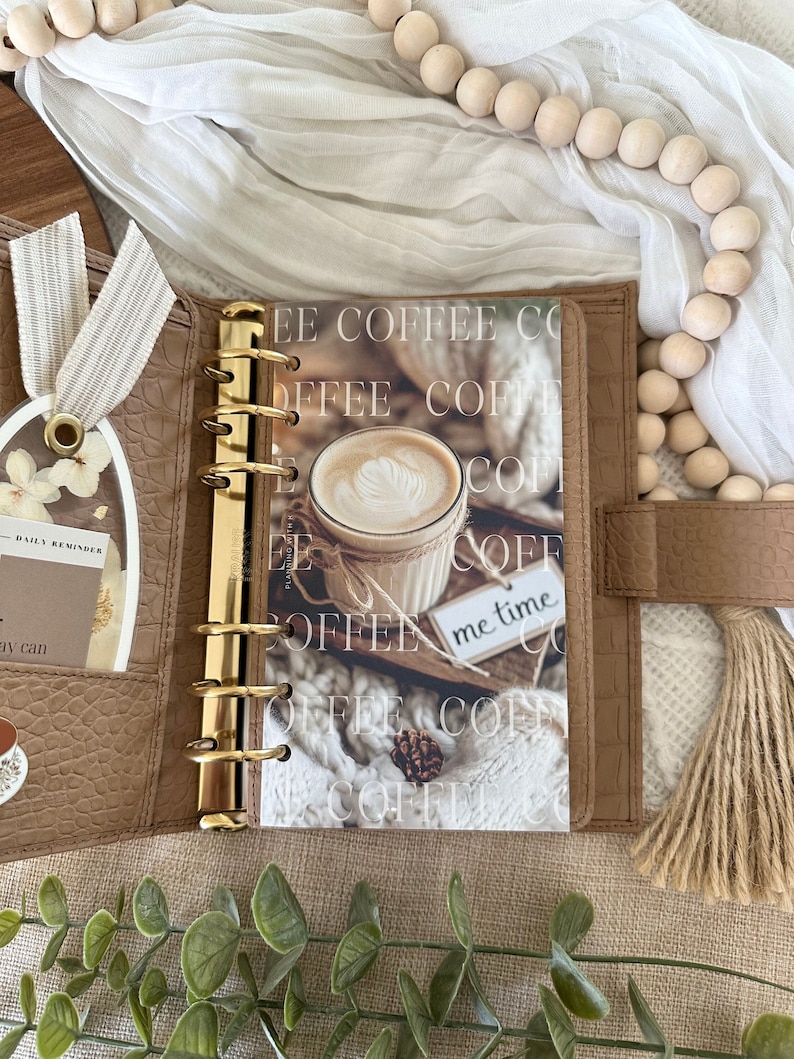 May include: A brown leather planner with gold rings and a page with a photo of a cup of coffee and the text "FEE COFFEE CO", "OFFEE COFE", "COFFEN", "COF", "COFF", "me time", "FEE COF"
