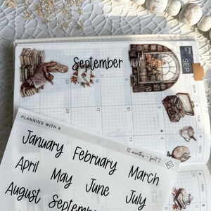 May include: A sheet of clear stickers with handwritten month names in black ink. The stickers are for use in a planner or journal. The month names are January, February, March, April, May, June, July, August, September, October, November, and December.