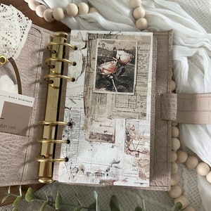 May include: A close-up of a light brown leather bound planner with gold rings. The planner is open to a page with a collage of vintage paper ephemera, including a black and white photo of a flower.
