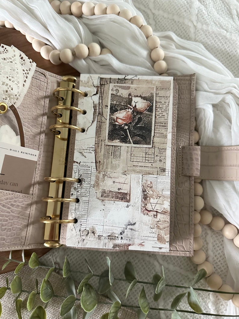 May include: A close-up of a light brown leather bound planner with gold rings. The planner is open to a page with a collage of vintage paper ephemera, including a black and white photo of a flower.
