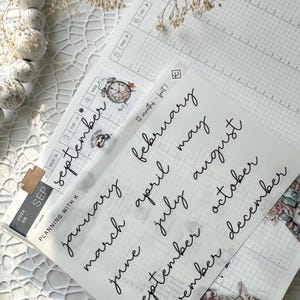 May include: A sheet of clear stickers with handwritten month names in black ink. The stickers are for use in a planner or journal.