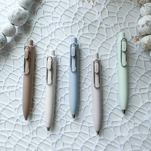 May include: Five pens in different colours: brown, white, light blue, light pink, and light green. The pens have a silver clip and a silver button on the top. The pens are arranged in a row on a white surface with a white lace pattern.