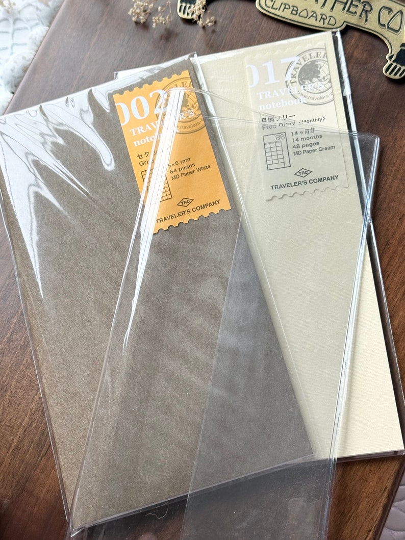 May include: Three Traveler's Company notebooks in brown paper covers with clear plastic wrapping. The notebooks are labeled "002 Traveler's Notebook", "017 Traveler's Notebook", and "Free Diary Monthly".