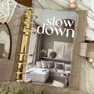 Puede incluir: Una carpeta dorada con un inserto de página de plástico transparente que presenta una foto de una sala de estar con un sofá seccional blanco y el texto "slow down".