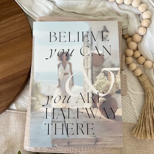 May include: A pink planner with the text "Believe you can & you are halfway there" on the cover. The cover features a blurry image of a person standing in front of a building.