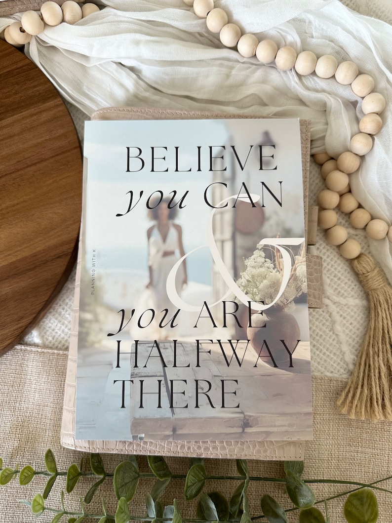 May include: A pink planner with the text "Believe you can & you are halfway there" on the cover. The cover features a blurry image of a person standing in front of a building.