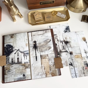 May include: An open journal with a brown leather cover and pages featuring a collage of vintage paper, a dragonfly, and a house illustration. A gold tray holds keychains with the phrases "Gratitude changes everything" and "Coffee & naps".