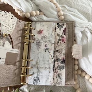 May include: A light beige leather bound planner with gold rings and a floral design on the inside page. The planner is open and the inside page has a white background with a floral design and a butterfly.