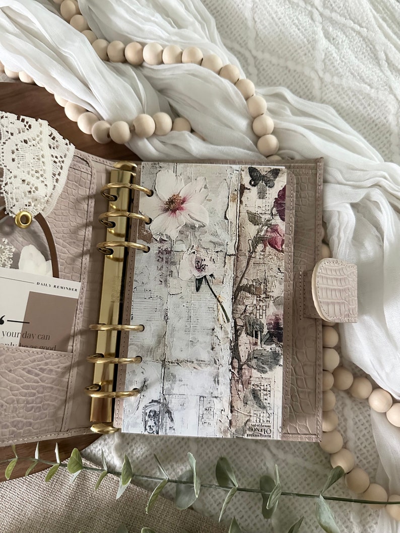 May include: A light beige leather bound planner with gold rings and a floral design on the inside page. The planner is open and the inside page has a white background with a floral design and a butterfly.