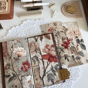 May include: A floral journal with a brown leather cover is open, displaying pages with a vintage rose design in red and cream. The journal is on a white lace doily, with pens and stationery in the background. A small tag reads "I just want to write."