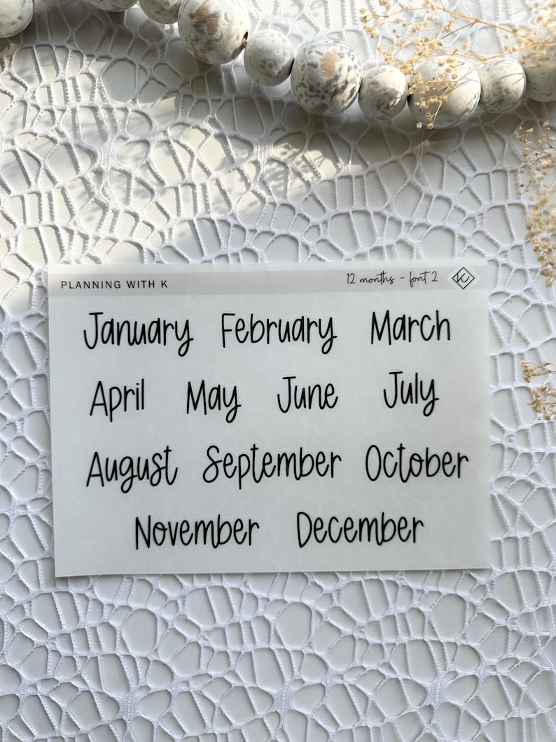 May include: A white sheet of paper with black text that reads "PLANNING WITH K" and "12 months - font 2". The sheet contains the months of the year written in a handwritten font.