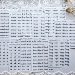 May include: A collection of white sticker sheets with black handwritten text. The stickers feature words like "anxious," "excited," and "happy." The brand name "Planning with K" is visible on some sheets. Decorative lace trim is at the bottom.