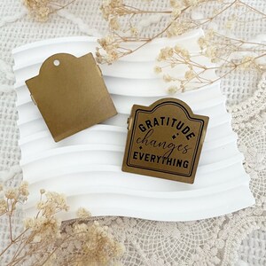 Puede incluir: Un sello de metal dorado con las palabras "Gratitude changes everything" en letras negras. El sello est&aacute; sobre una superficie blanca con un patr&oacute;n decorativo.