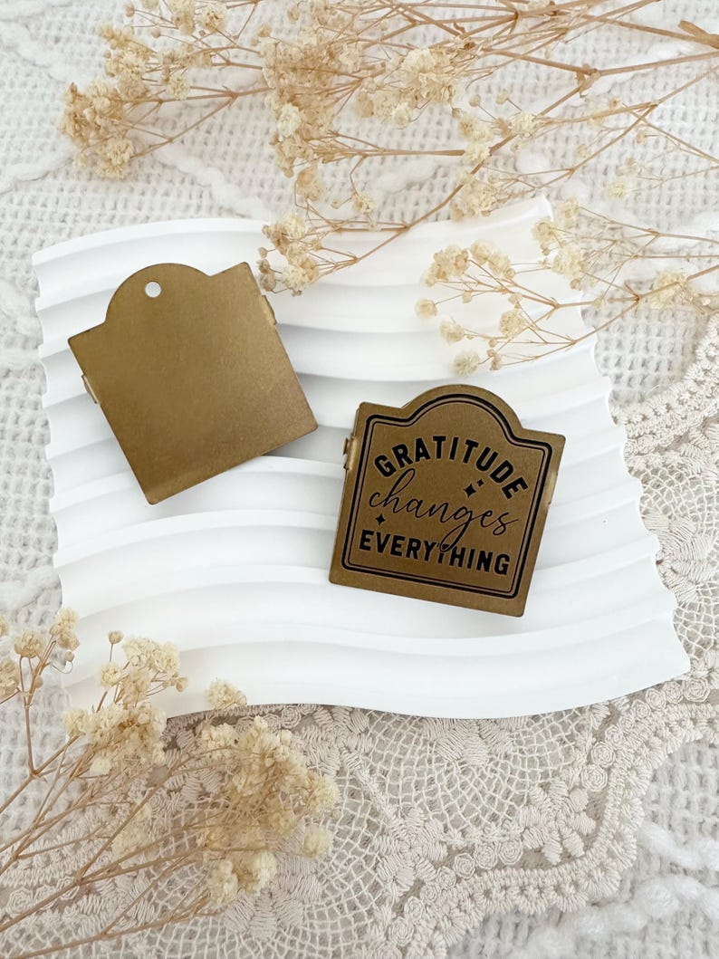Puede incluir: Un sello de metal dorado con las palabras "Gratitude changes everything" en letras negras. El sello est&aacute; sobre una superficie blanca con un patr&oacute;n decorativo.