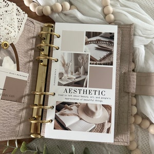 Pode incluir: Uma agenda branca com argolas douradas e uma p&aacute;gina que diz "Est&eacute;tica" com uma defini&ccedil;&atilde;o da palavra. A p&aacute;gina tem v&aacute;rias imagens de um livro, um chap&eacute;u, um vaso com flores secas e um caderno com l&aacute;pis.