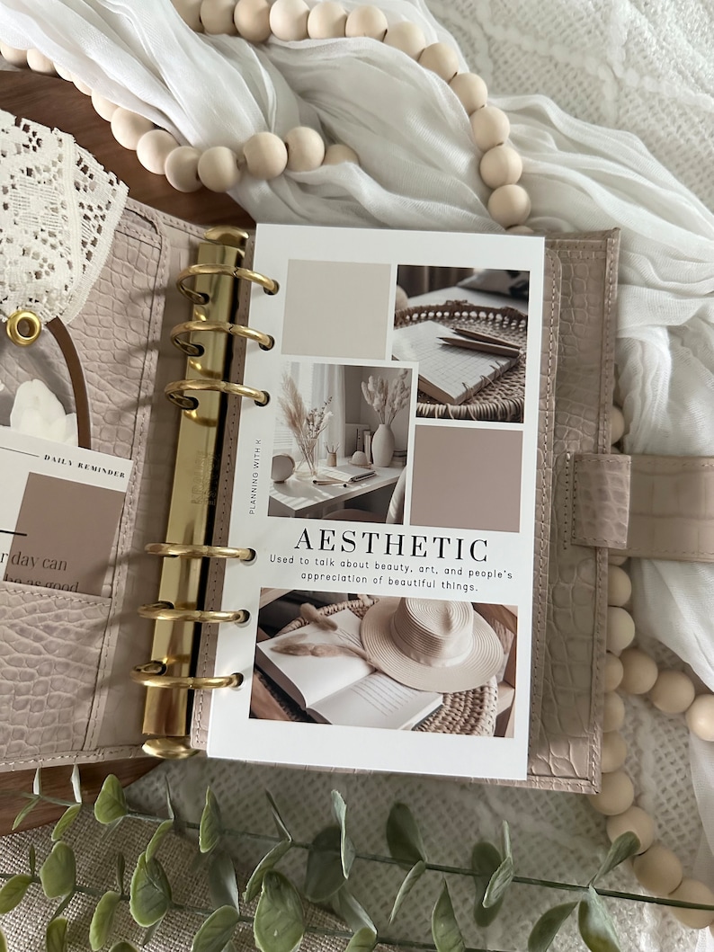Pode incluir: Uma agenda branca com argolas douradas e uma p&aacute;gina que diz "Est&eacute;tica" com uma defini&ccedil;&atilde;o da palavra. A p&aacute;gina tem v&aacute;rias imagens de um livro, um chap&eacute;u, um vaso com flores secas e um caderno com l&aacute;pis.