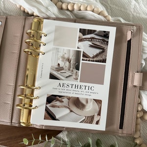 Pode incluir: Uma agenda branca com argolas douradas e uma p&aacute;gina que diz "AESTHETIC" com uma defini&ccedil;&atilde;o da palavra. A p&aacute;gina tem imagens de estilo colagem de um livro, um chap&eacute;u, um vaso com flores e um caderno com l&aacute;pis.
