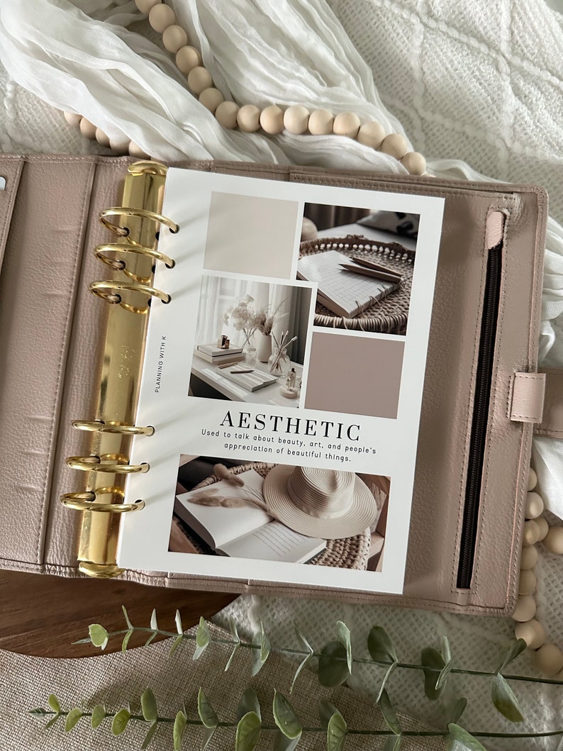 Pode incluir: Uma agenda branca com argolas douradas e uma p&aacute;gina que diz "AESTHETIC" com uma defini&ccedil;&atilde;o da palavra. A p&aacute;gina tem imagens de estilo colagem de um livro, um chap&eacute;u, um vaso com flores e um caderno com l&aacute;pis.