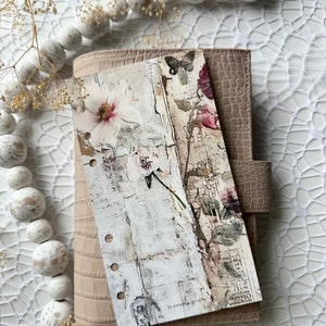 May include: A beige leather planner with a floral design bookmark. The bookmark features a white flower with pink accents and a butterfly. The planner has a textured, crocodile-embossed cover.