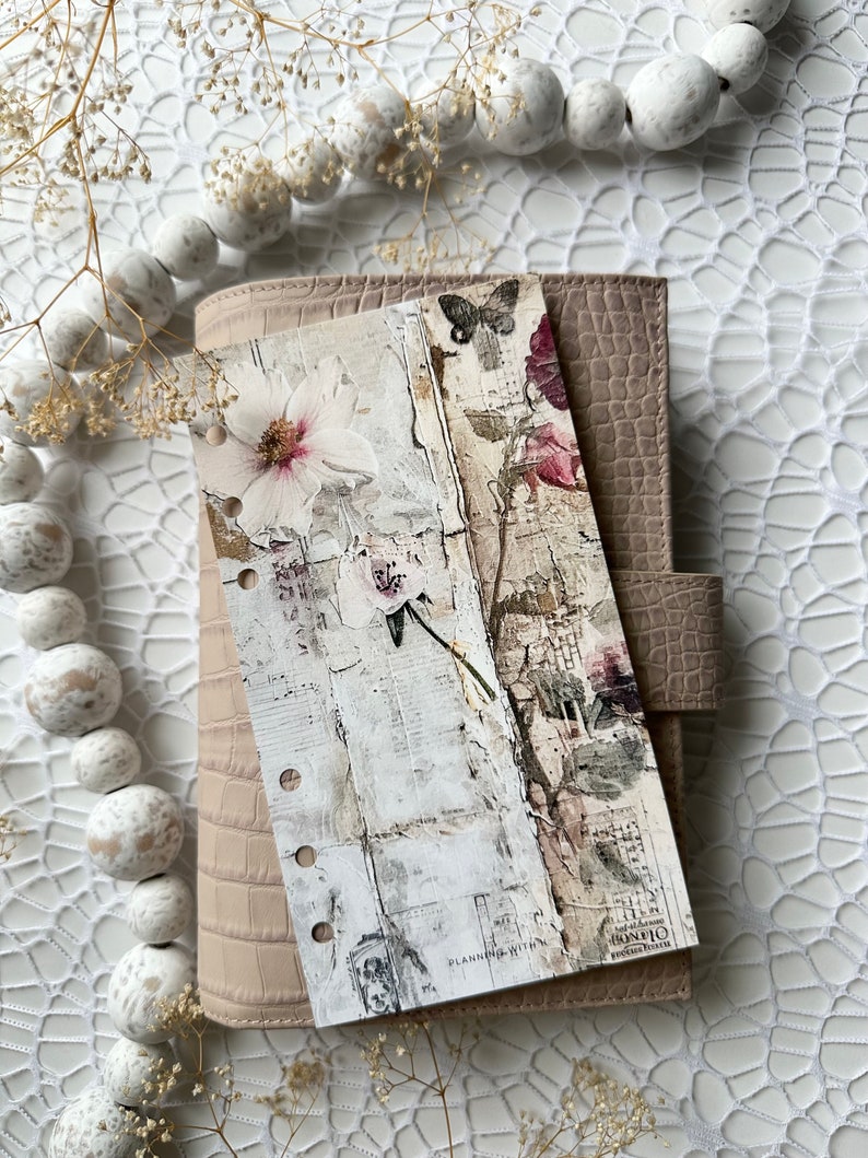 May include: A beige leather planner with a floral design bookmark. The bookmark features a white flower with pink accents and a butterfly. The planner has a textured, crocodile-embossed cover.