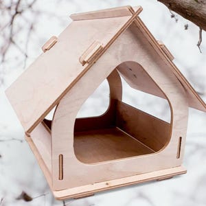 Op de afbeelding: Een houten vogelvoederhuisje in de vorm van een huis met een schuin dak en een grote opening aan de voorkant. De voederbak is gemaakt van licht hout en heeft een natuurlijke afwerking.