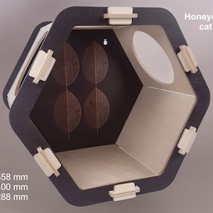 Puede incluir: Una estantería hexagonal para gatos en forma de panal hecha de madera con un acabado negro y beige. La estantería tiene una abertura redonda y un patrón de hojas decorativo en la parte posterior. La estantería mide 358 mm de alto, 400 mm de ancho y 288 mm de profundidad.