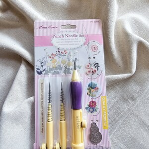 May include: A pink and yellow packaging for a punch needle set. The set includes three yellow punch needles with purple handles and a small metal tool. The packaging has the text "Mina Carin Punch Needle Set" and "Quality Product".