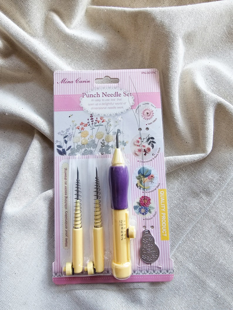 May include: A pink and yellow packaging for a punch needle set. The set includes three yellow punch needles with purple handles and a small metal tool. The packaging has the text "Mina Carin Punch Needle Set" and "Quality Product".