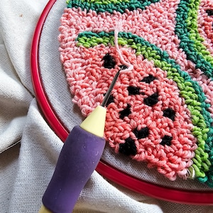May include: A close-up of a pink and green watermelon-themed punch needle project in progress. The needle is inserted into the fabric, and the yarn is being pulled through. The project is being worked on in a hoop.