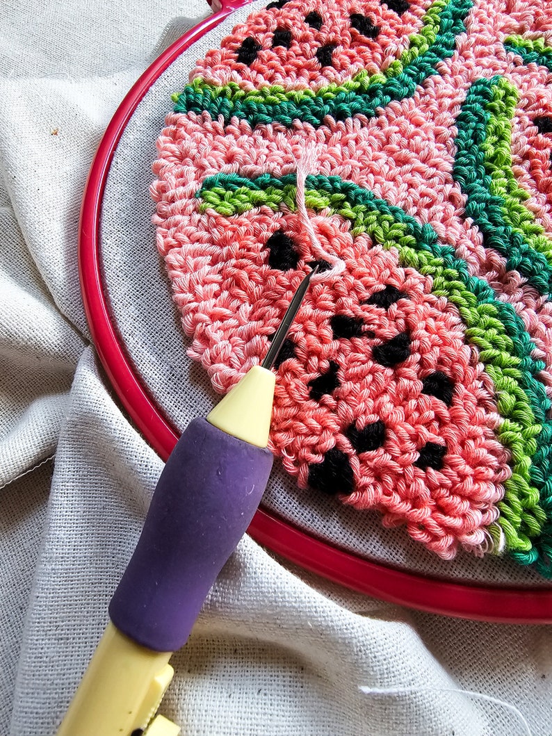 May include: A close-up of a pink and green watermelon-themed punch needle project in progress. The needle is inserted into the fabric, and the yarn is being pulled through. The project is being worked on in a hoop.