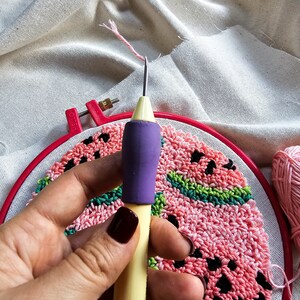 May include: A close-up of a hand holding a yellow and purple punch needle tool. The needle is inserted into a red embroidery hoop with a pink and green watermelon design.