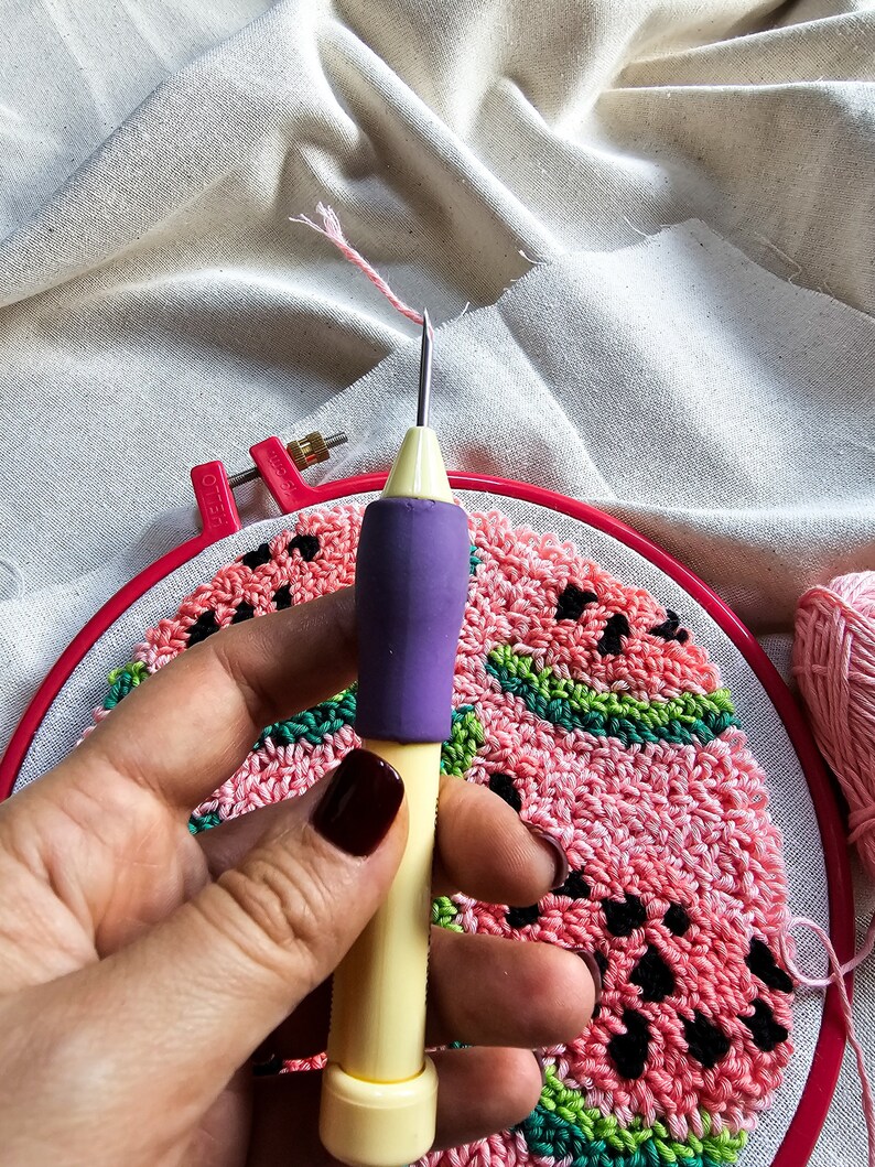 May include: A close-up of a hand holding a yellow and purple punch needle tool. The needle is inserted into a red embroidery hoop with a pink and green watermelon design.