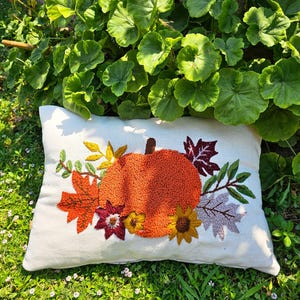 K&ouml;nnte beinhalten: Ein wei&szlig;es Kissen mit einer gestickten K&uuml;rbis, Bl&auml;ttern und Blumen in Orange-, Gelb-, Rot- und Lilat&ouml;nen. Das Kissen liegt auf einem Bett aus gr&uuml;nem Gras.