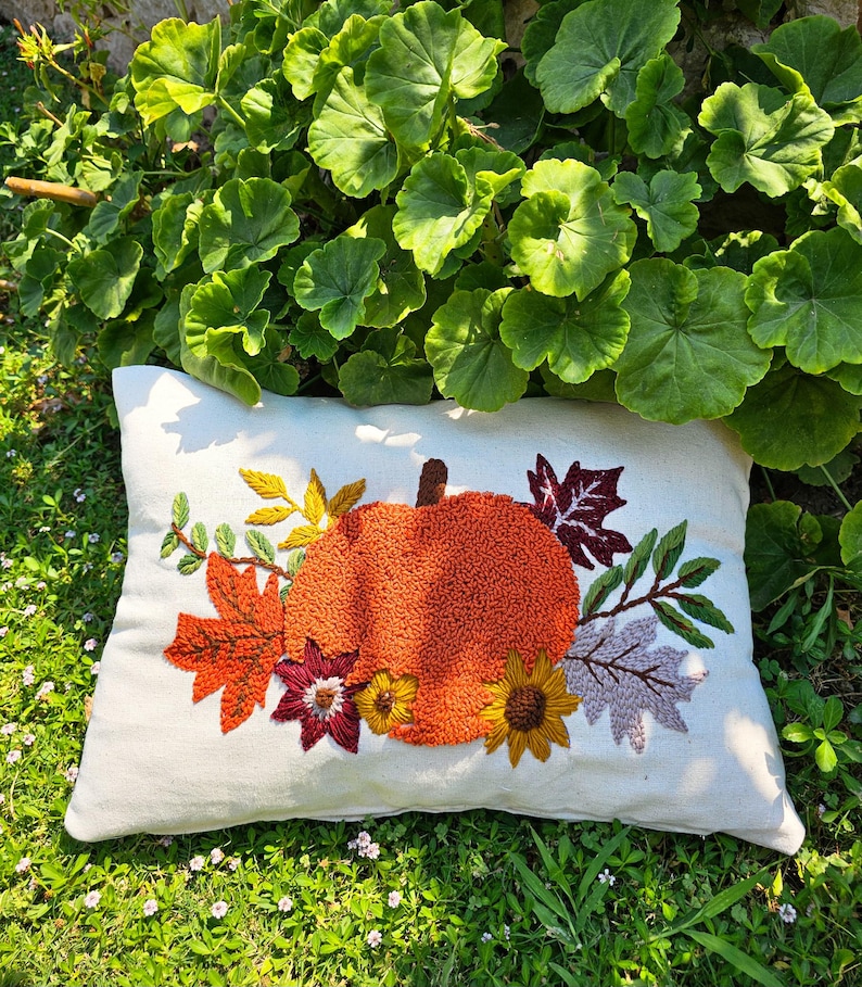 K&ouml;nnte beinhalten: Ein wei&szlig;es Kissen mit einer gestickten K&uuml;rbis, Bl&auml;ttern und Blumen in Orange-, Gelb-, Rot- und Lilat&ouml;nen. Das Kissen liegt auf einem Bett aus gr&uuml;nem Gras.