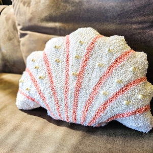 Puede incluir: Una almohada con forma de concha de mar de color blanco y rosa con una superficie texturizada y detalles de perlas blancas.