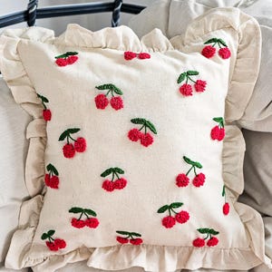 Puede incluir: Cojín cuadrado color crema con cerezas rojas bordadas y hojas verdes. El cojín tiene un borde con volantes y está colocado sobre una cama. Las cerezas están distribuidas uniformemente por la superficie del cojín.