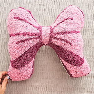 Peut inclure: Un coussin décoratif rose en forme de nœud papillon avec un design texturé et touffeté. Le nœud présente une base rose clair avec des rayures et des accents roses plus foncés. Le coussin est sur une surface de ton neutre.