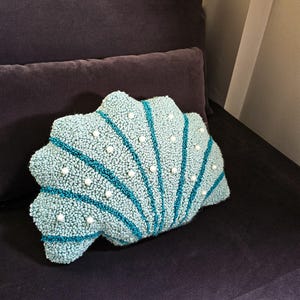 May include: A teal blue and white seashell-shaped cushion with a textured surface and white pearl accents.