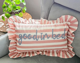 May include: A pink and white striped pillow with a ruffled edge. The pillow has the words "good in bed" embroidered in blue thread.