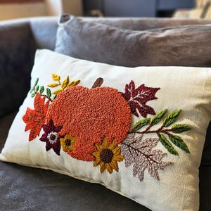 K&ouml;nnte beinhalten: Ein wei&szlig;es Kissen mit einer gestickten K&uuml;rbis, Bl&auml;ttern und Blumen in Orange-, Rot-, Gelb-, Gr&uuml;n- und Lilat&ouml;nen. Das Kissen hat einen Rei&szlig;verschluss.