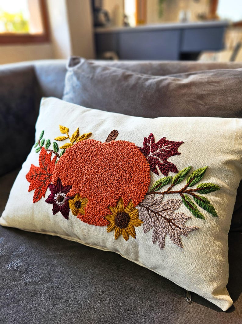 K&ouml;nnte beinhalten: Ein wei&szlig;es Kissen mit einer gestickten K&uuml;rbis, Bl&auml;ttern und Blumen in Orange-, Rot-, Gelb-, Gr&uuml;n- und Lilat&ouml;nen. Das Kissen hat einen Rei&szlig;verschluss.