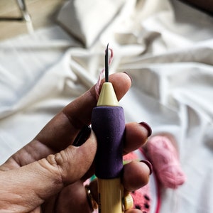 May include: A close-up of a hand holding a yellow and purple embroidery needle threader. The needle is threaded with pink thread. The needle threader is on a white fabric background.