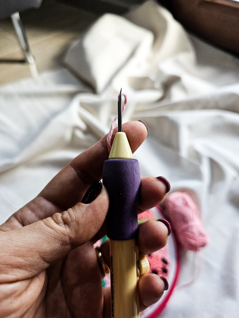 May include: A close-up of a hand holding a yellow and purple embroidery needle threader. The needle is threaded with pink thread. The needle threader is on a white fabric background.