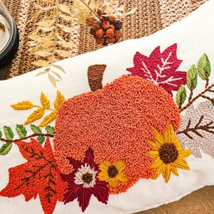 K&ouml;nnte beinhalten: Ein wei&szlig;es Kissen mit einer gestickten K&uuml;rbis, Bl&auml;ttern und Blumen in Orange-, Rot-, Gelb- und Gr&uuml;nt&ouml;nen. Das Kissen ist perfekt f&uuml;r die Herbstdekoration.