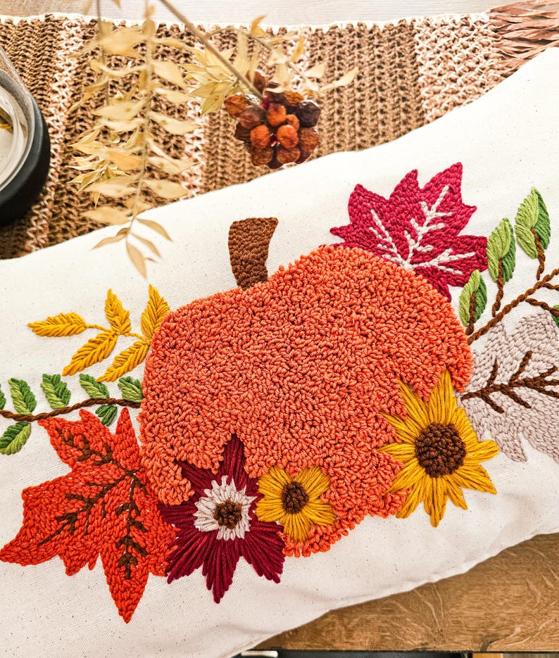 K&ouml;nnte beinhalten: Ein wei&szlig;es Kissen mit einer gestickten K&uuml;rbis, Bl&auml;ttern und Blumen in Orange-, Rot-, Gelb- und Gr&uuml;nt&ouml;nen. Das Kissen ist perfekt f&uuml;r die Herbstdekoration.